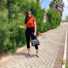 a woman in black pants and an orange top is standing on a brick road with trees behind her