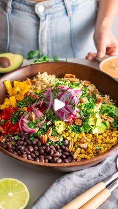 a woman holding a bowl filled with vegetables and beans on top of a table next to sliced avocado