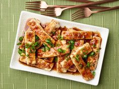 tofu with sauce and green onions on a plate next to utensils, fork and spoons