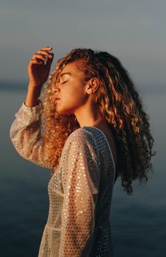 a woman with curly hair standing by the water holding her hand up to her ear