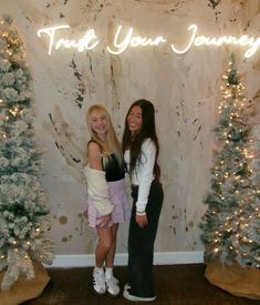 two girls standing next to each other in front of a wall with christmas trees and lights