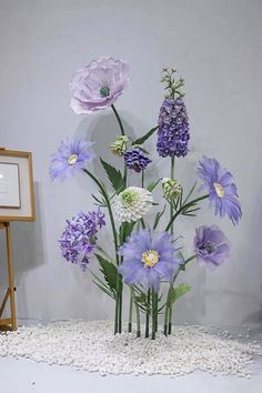 an arrangement of purple and white flowers is displayed in front of a gray wall with pebbles