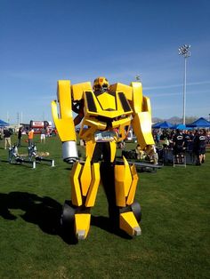 a large yellow and black robot standing on top of a lush green field next to people