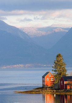 a red house sitting on the shore of a lake with mountains in the back ground