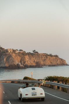 a white car driving down the road next to a hill and ocean with a cliff in the background