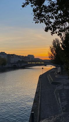 the sun is setting over the water and people are walking on the sidewalk near the river