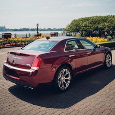 a red car parked on the side of a road next to flowers and trees in front of a body of water