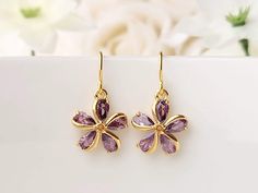 Purple gemstone petals, are the highlight of these dainty, floral earrings. They are made from genuine zircon stones, secured inside an 18k gold plated setting. They radiate a lovely, sparkly shine! T Product details: * Gold plated ear wires  * Lead and nickel free * 18k gold plated setting * Zircon gemstones -  Length:  .8 inches -  Width:    .5 inches ❤ I also have these earrings available in emerald green, multi-colored, pink, and clear, in my shop.  https://www.etsy.com/shop/FlutterbirdCo My goal is to provide every customer with exceptional service. Please message me if you have any questions. Thank you for supporting my small business!  Please visit my F.A.Q. and shop policy sections for additional info. Purple Minimalist, Zircon Gemstone, Gold And Purple, Gemstone Earrings Gold, Flower Dangle Earrings, Purple Flower, Floral Earrings, Star Earrings, Heart Earrings