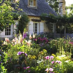 a house surrounded by flowers and greenery in front of the building with white windows