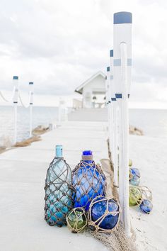 blue and green glass bottles sitting on the sand next to an ocean shore with a white house in the background