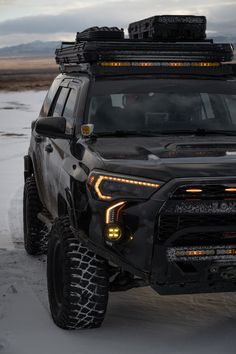 the front end of a black truck with its lights on in the middle of snow