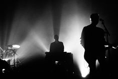 the silhouettes of two people on stage with lights behind them and drums in front of them