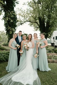 a group of people standing next to each other in front of trees and grass with one woman wearing a wedding dress