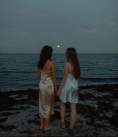 two women standing on the beach holding hands and looking out at the ocean under a full moon