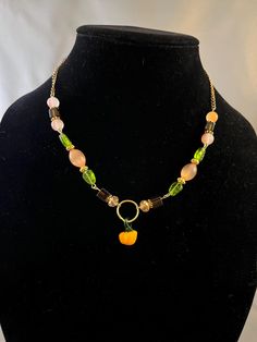 a gold tone necklace decorated with orange and brown beads with green leaf beads and gold tone spacers. A gold tone hoop connects the two sides of the necklace with a small pumpkin hanging from the bottom of the hoop. Pumpkin Necklace, Leaf Beads, Orange Decor, Small Pumpkins, Gold Tone Necklace, Green Leaf, Fall Pumpkins, Green Leaves, Gold Tones