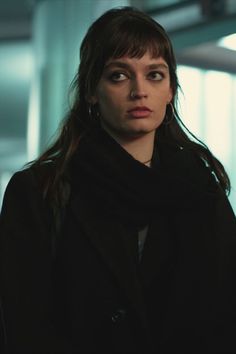 a woman with long hair wearing a black coat and looking at the camera while standing in an empty room