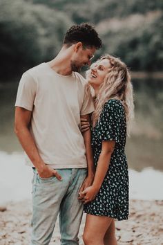 a young man and woman standing next to each other on the shore of a lake