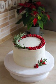 a white cake with red berries and greenery on it sitting on a plate next to a potted plant