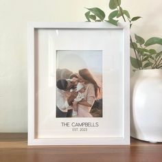 a white vase with a plant in it next to a photo frame that says the campbells