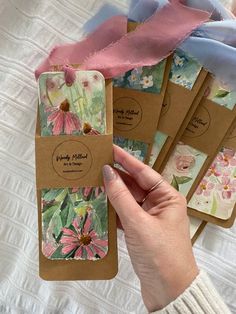 the hand is holding four small tags with flowers on them, and one has a pink ribbon around it