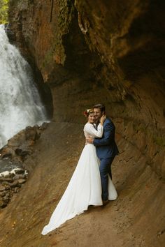 a bride and groom standing in front of a waterfall at the base of a cliff