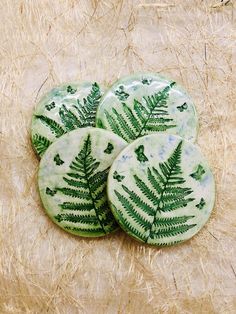 three green buttons with leaves on them sitting on some straw covered ground and one has a fern leaf painted on it