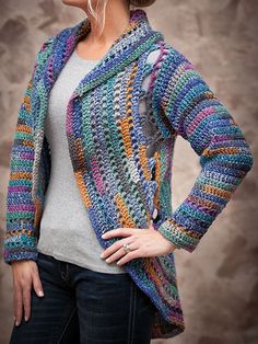 a woman wearing a multicolored crocheted jacket and jeans standing in front of a wall