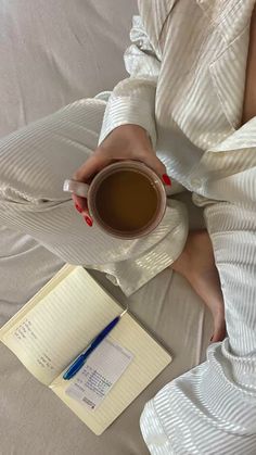 a woman is sitting on her bed holding a cup of coffee and an open notebook