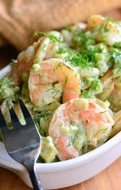 a white bowl filled with shrimp and pasta salad next to a fork on top of a wooden table