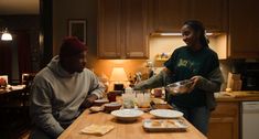 two people sitting at a kitchen table with plates of food on it and one person holding a bowl