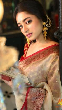 a woman in a red and gold sari holding a book while standing next to a lamp