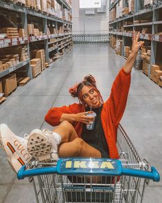 a woman sitting on top of a shopping cart in a warehouse holding a drink and waving