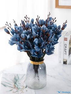 blue flowers are in a glass vase on a marble countertop next to a book