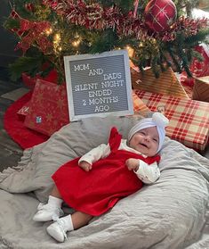 a baby in a red dress laying on top of a bed next to a christmas tree