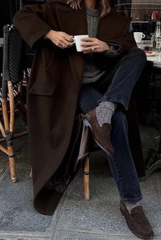 a woman sitting in a chair holding a cup and looking at her cell phone while wearing a brown coat