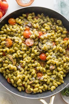 pasta with pesto and tomatoes in a skillet on a white table next to other ingredients