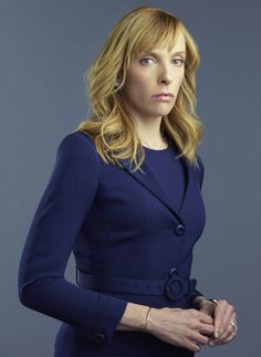 a woman with blonde hair wearing a blue coat and bracelets, standing in front of a gray background