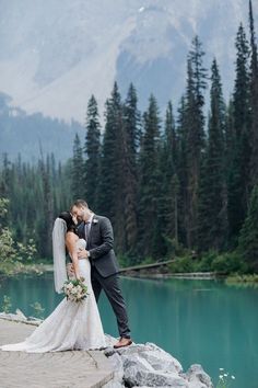 a bride and groom kissing on the edge of a cliff overlooking a lake surrounded by pine trees