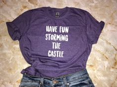 "A fun, and soft shirt for fans of The Princess Bride. This comfy shirt makes an awesome gift and goes well with everything from jeans and boots to leggings and sneakers. *Processing time is currently 4-9 business days. Thank you for your patience!* Design: \"Have Fun Storming The Castle\" Shirt Sizing Softstyle (Unisex Adult T-Shirt) - Comfortable for both women and men to wear. - Please see size chart in the photos for more details. Materials - 100% preshrunk ringspun cotton - Heather colors 3 Funny Text Tops For Fan Merchandise, Casual Purple T-shirt For Fan Merchandise, Casual Purple T-shirt For Fans, Casual Purple Slogan T-shirt, Casual All-gender Slogan Shirt, Unisex Casual Slogan Shirt, Fun Slogan T-shirt For Fan Merchandise, Funny Text Graphic Tee Fan Merchandise, Casual Pre-shrunk Shirt For Fan Conventions