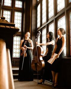 three women standing in front of stained glass windows, one holding a cello and the other wearing a black dress