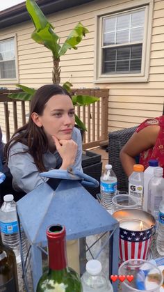 a woman sitting at a table with bottles and glasses on it in front of a house