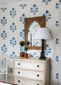 a dresser with a lamp and mirror on it in front of a blue flowered wallpaper