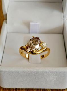 an engagement ring in a white box on a wooden table with a gold band and brown diamond