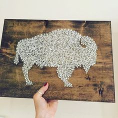 a person holding up a string art bison on a wooden board with white thread in the shape of a buffalo