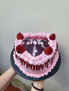 someone holding a cake with pink icing and strawberries on top, in front of a wall