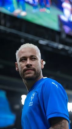 a man with white hair wearing a blue shirt in front of a large television screen