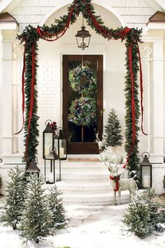 a white house decorated for christmas with wreaths and garland on the front door, lanterns and trees