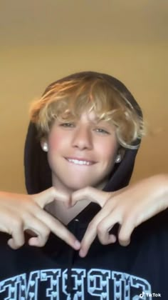 a young boy with blonde hair wearing a blue shirt and holding his hands in the shape of a heart