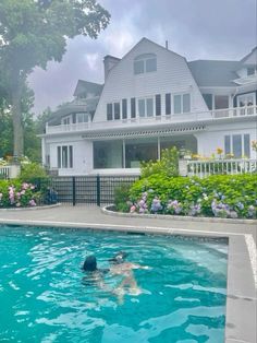 a person swimming in a pool next to a large white house with flowers around it