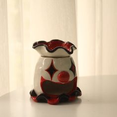 a red and white vase sitting on top of a table next to a window curtain